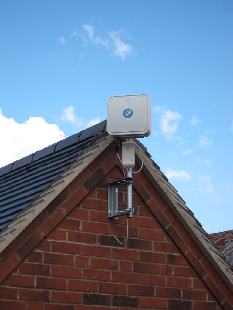 A colour photograph showing the top part of the red-brick gable wall of a building and part of a grey tile roof. The main focus of the photograph is a square white plastic enclosure with a blue circular label in the centre, mounted on a vertical aluminium pole attached to the wall with a silver-painted metal bracket.
Blue sky with a few clouds is visible in the background.